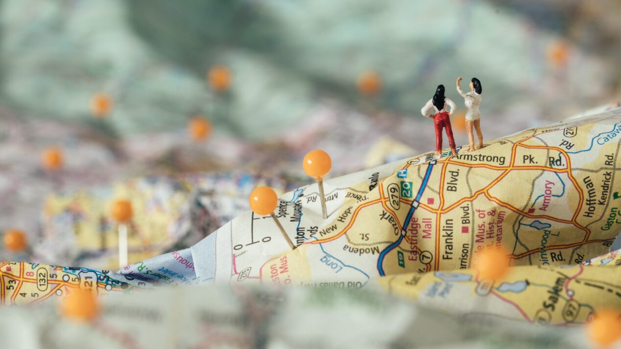 A diorama-like image of a 3D paper map with yellow pins stuck in it and two tiny human figures standing on one ridge.