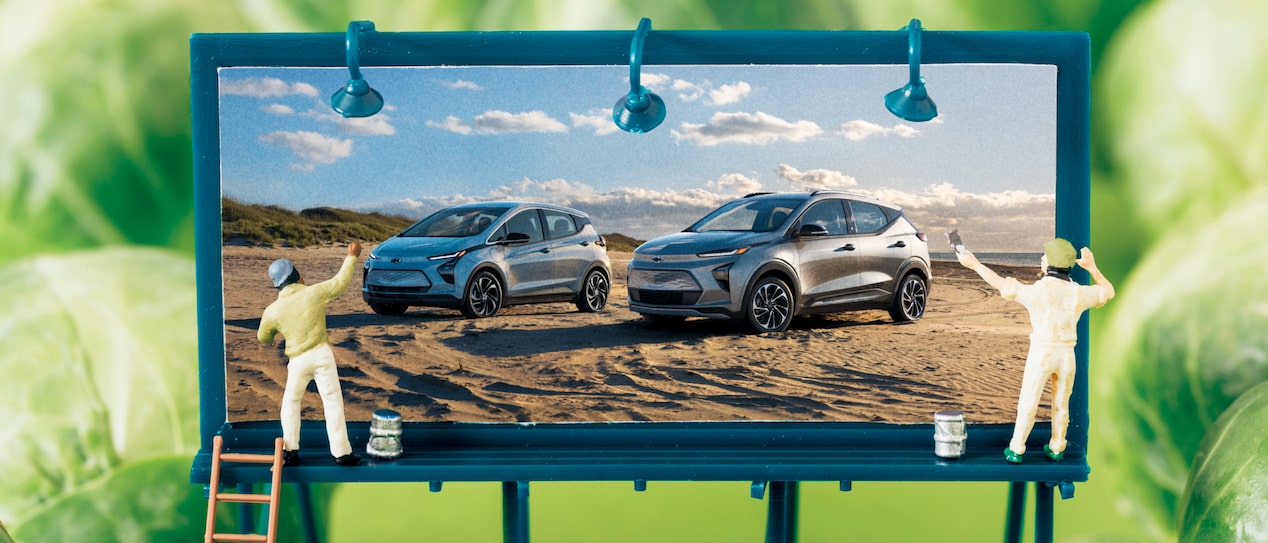 A diorama-like image of a miniature billboard showing the Bolt EV and Bolt EUV, with two tiny human figures painting it, surrounded by Brussels sprouts.