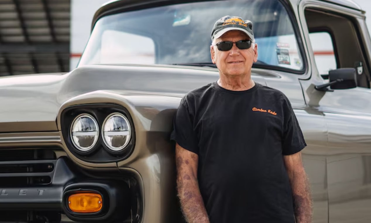 Un hombre con una camiseta negra y anteojos de sol posando en frente de una pickup Chevrolet clásica